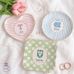 May include: Three decorative ceramic trays with gold rims. One is heart-shaped with pink stripes and the name "Ava" and "Class of 2026". Another is round with blue stripes and the name "Olivia" and "Class of 2026". The third is square with a green checkered pattern and the name "Isabella" and "Class of 2026".