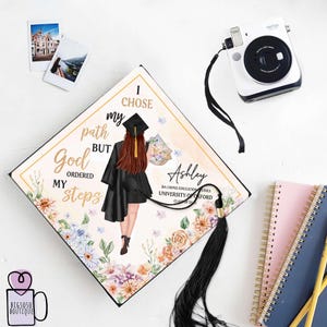 May include: A graduation cap with a floral design and a black tassel. The cap features a quote that reads "I chose my path but God ordered my steps." The cap also includes the graduate's name, Ashley, and their degree information.