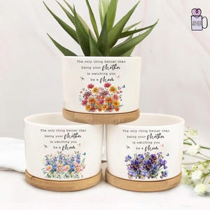 May include: Three white ceramic flower pots with wooden saucers, each adorned with floral designs and the text "The only thing better than being your Mother is watching you be a Mom." One pot holds a green plant.