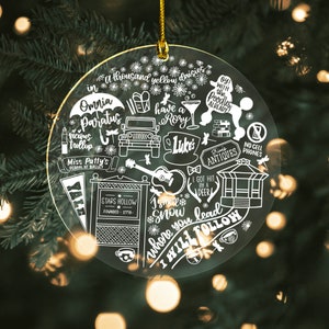 May include: A clear round Christmas ornament with white illustrations of scenes and quotes from the TV show Gilmore Girls. The ornament features text such as "Omnia Paratus", "Stars Hollow", "I smell snow", "Luke's", "Where you lead I will follow", and "Kim's Antiques".