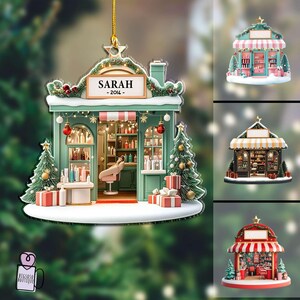 May include: A Christmas ornament shaped like a beauty salon, with a sign that reads "SARAH - 2014-". The salon is painted in shades of green and pink, with a red and white striped awning. The ornament is decorated with Christmas trees, presents, and lights.