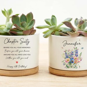 May include: Two white ceramic planters with succulents. One planter reads "Chapter Sixty" with a birthday message. The other planter is personalized with the name "Jennifer" and floral artwork. Both planters sit on wooden saucers.
