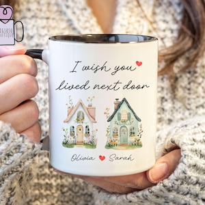 Può includere: Tazza in ceramica bianca con bordo e manico neri. La tazza presenta il testo "I wish you lived next door" con due case illustrate e i nomi "Olivia" e "Sarah". È incluso un simbolo a forma di cuore.