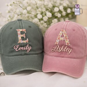 May include: Two baseball caps, one olive green and one dusty rose, are displayed. The caps feature floral letter designs and embroidered names: "E" and "Emily" on the green cap, and "A" and "Ashley" on the pink cap. White flowers are in the background.
