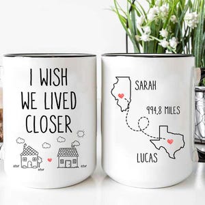 May include: Two white coffee mugs with black handles. One mug says "I wish we lived closer" with a house illustration. The other mug says "Sarah 994.8 miles Lucas" with a map illustration of Illinois and Texas.