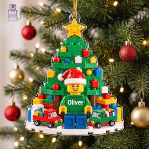 May include: A flat, clear acrylic Christmas tree ornament shaped like a pine tree built from green building blocks. It features a yellow star topper, a central minifigure wearing a Santa hat and a green sweater with the name Oliver printed on it.