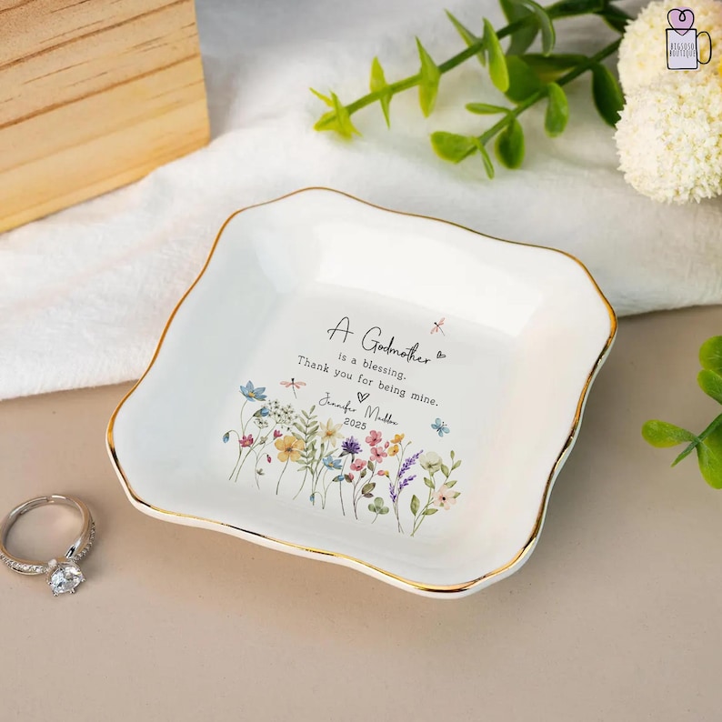 Pu&ograve; includere: Piatto portaanelli in ceramica bianca con bordo dorato e design floreale. Il piatto presenta la scritta "A Godmother is a blessing. Thank you for being mine." e un nome e un anno. Un anello d'argento &egrave; in primo piano.