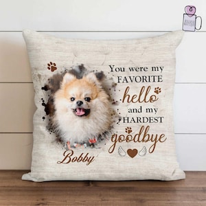 May include: A beige square pillow featuring a Pomeranian dog portrait with the name "Bobby". The text reads "You were my FAVORITE hello and my HARDEST goodbye" in brown script. Paw prints and a heart are also included.