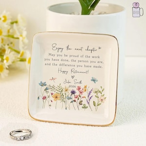 May include: A square, white ceramic trinket dish with a gold rim, featuring a floral design and the text "Enjoy the next chapter." The dish includes the text "Happy Retirement!" and a name and year. A silver ring with clear stones sits in front.