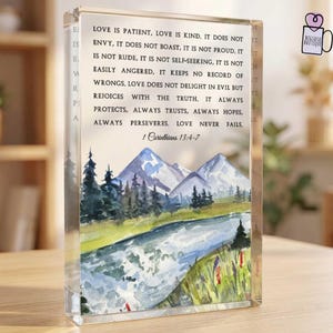 May include: A rectangular clear acrylic block featuring a watercolor landscape of a river, pine trees, and mountains. The text reads: Love is patient, love is kind. It does not envy, it does not boast, it is not proud. It is not rude, it is not self-seeking, it is not easily angered, it keeps no record of wrongs. Love does not delight in evil but rejoices with the truth. It always protects, always trusts, always hopes, always perseveres. Love never fails. 1 Corinthians 13:4-7.