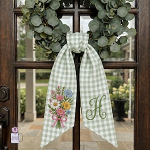 May include: A green and white gingham ribbon with a floral bouquet embroidery and the letter "H" on a wooden door. The ribbon is tied around a eucalyptus wreath. The bouquet features pink, blue, and yellow flowers.