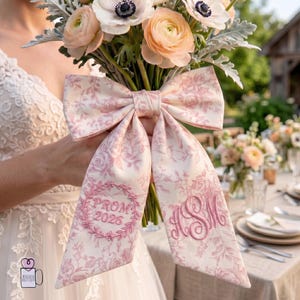 May include: A large, pink floral bow with the text "PROM 2026" and monogrammed initials. The bow is tied around a bouquet of flowers with peach and white blooms. The bow has a floral pattern.