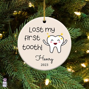 Pu&ograve; includere: Un addobbo natalizio in ceramica bianca con un design a forma di dente di cartone animato. L'addobbo dice "Lost my first tooth!" e ha il nome "Henry" e l'anno "2023" stampati su di esso.