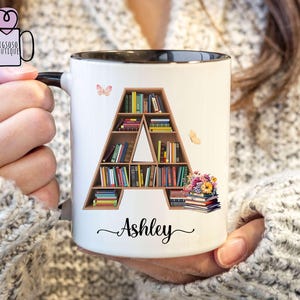 May include: White ceramic mug with a black rim. The mug features a bookshelf shaped like the letter "A" filled with books. The name "Ashley" is written below the bookshelf.