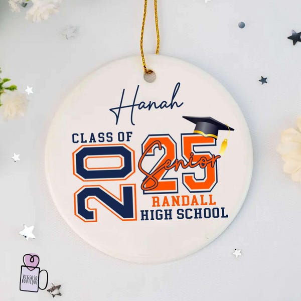 Pode incluir: Ornamento de cerâmica branca com o texto "Class of 2025 Senior" em letras laranja e azul. O texto "Hannah" está escrito em itálico no topo do ornamento. O texto "Randall High School" está escrito abaixo do ano. Um chapéu de formatura é representado acima do ano.