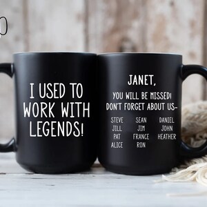K&ouml;nnte beinhalten: Zwei schwarze Keramikbecher. Auf einem Becher steht in wei&szlig;er Schrift "I USED TO WORK WITH LEGENDS!". Der andere Becher tr&auml;gt die Aufschrift "JANET, YOU WILL BE MISSED! DON'T FORGET ABOUT US-" mit einer Namensliste. Die Becher haben schwarze Griffe.