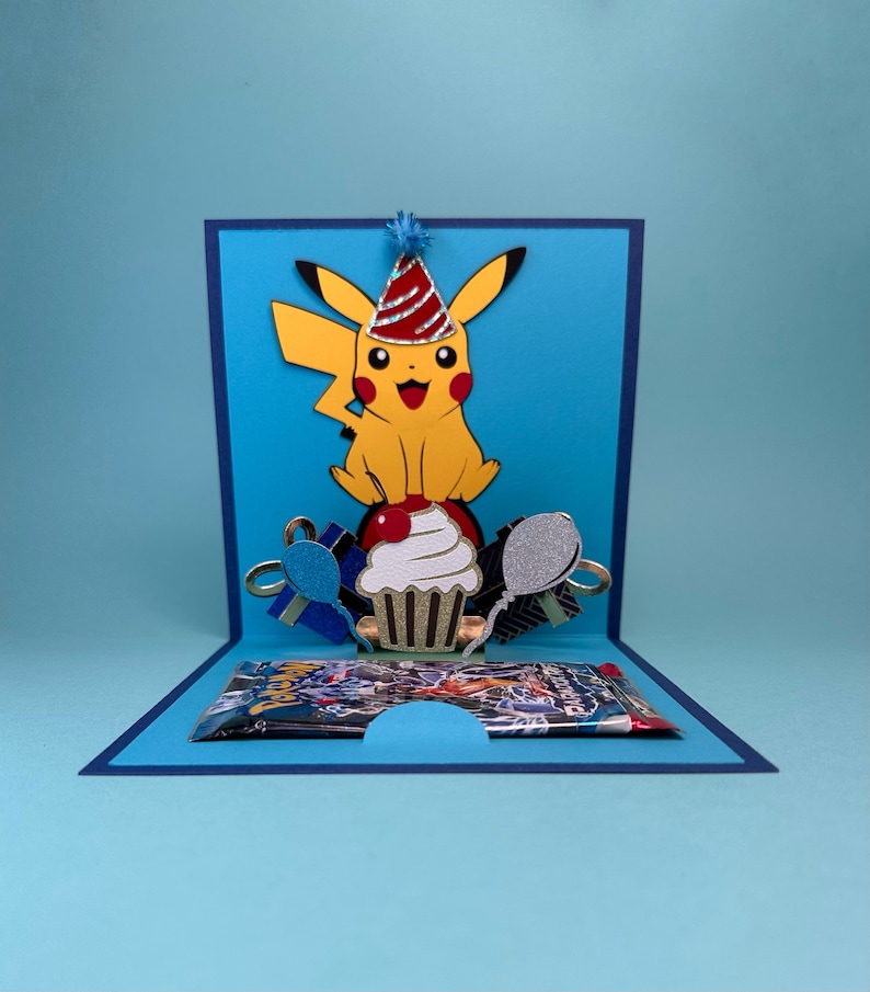 May include: A handmade pop-up birthday card featuring a yellow Pikachu character wearing a party hat. The card includes a cupcake, balloons, and gift boxes. The card is blue with a dark blue border and a gift card inside.