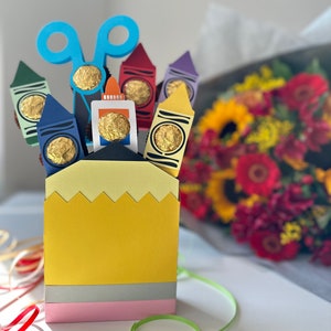 May include: A pencil-shaped gift box with a yellow top, gray middle, and pink base. It holds crayon-shaped cutouts with gold-wrapped candies. Blue scissors and a glue bottle cutout are also included. A bouquet of flowers is in the background.