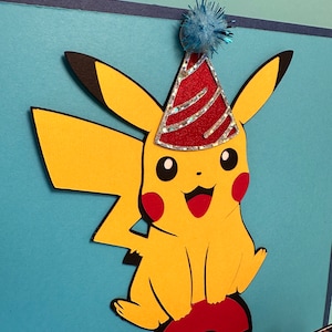May include: A yellow Pikachu paper cutout wearing a red and silver party hat, perched on a red and white Poke Ball. The background is a light blue with a dark blue border. A blue pom-pom is attached to the top of the hat.