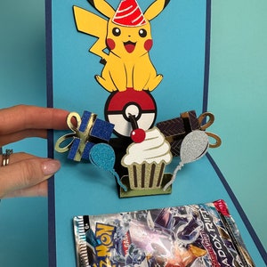 May include: A handmade birthday card featuring a yellow Pikachu character wearing a party hat, atop a Poke Ball. The card includes a cupcake, gifts, and balloons. A Pokemon card pack is visible in the card's pocket. The card is blue with dark blue accents.