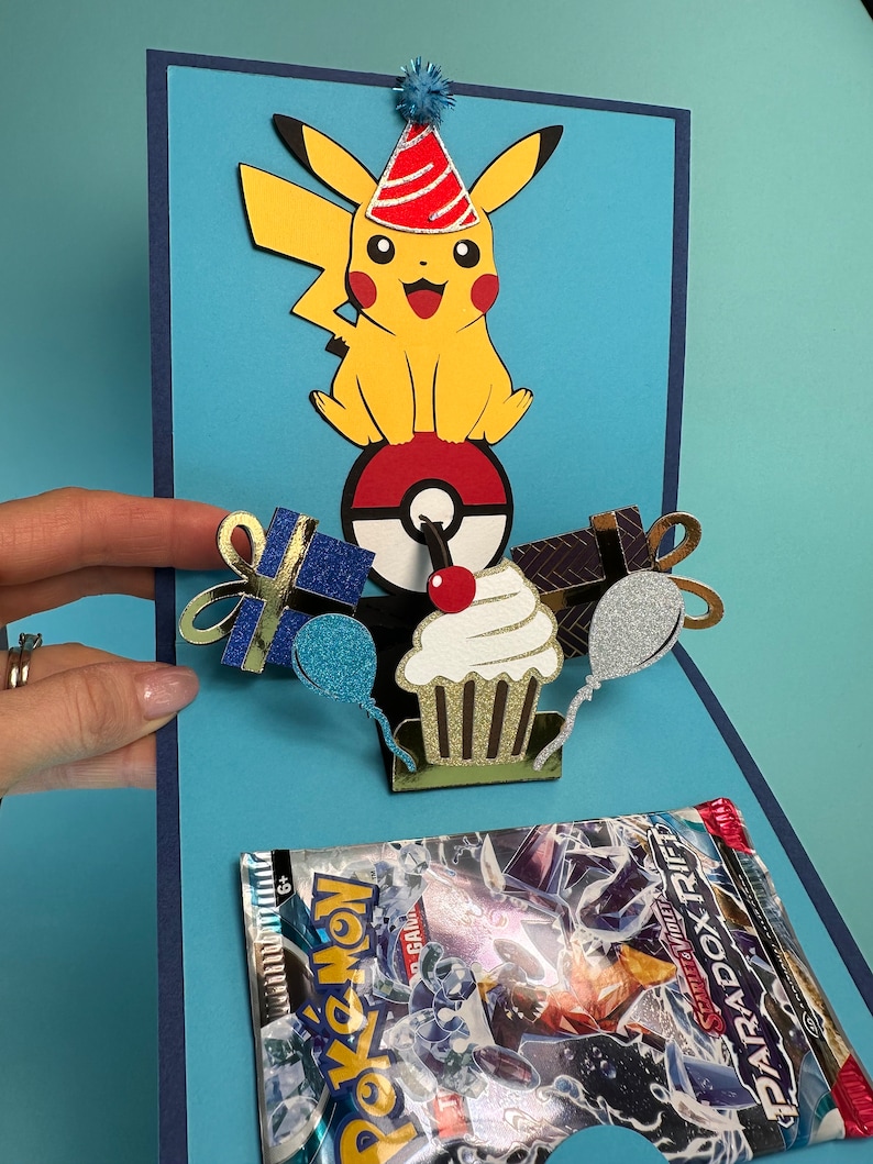 May include: A handmade birthday card featuring a yellow Pikachu character wearing a party hat, atop a Poke Ball. The card includes a cupcake, gifts, and balloons. A Pokemon card pack is visible in the card's pocket. The card is blue with dark blue accents.