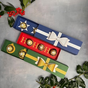 Può includere: Quattro scatole regalo rettangolari blu, rosse e verdi, ciascuna contenente cioccolatini. La scatola blu ha un fiocco argentato e decorazioni a fiocco di neve. La scatola rossa dice "HO HO HO" in oro. La scatola verde ha un fiocco dorato e ornamenti.