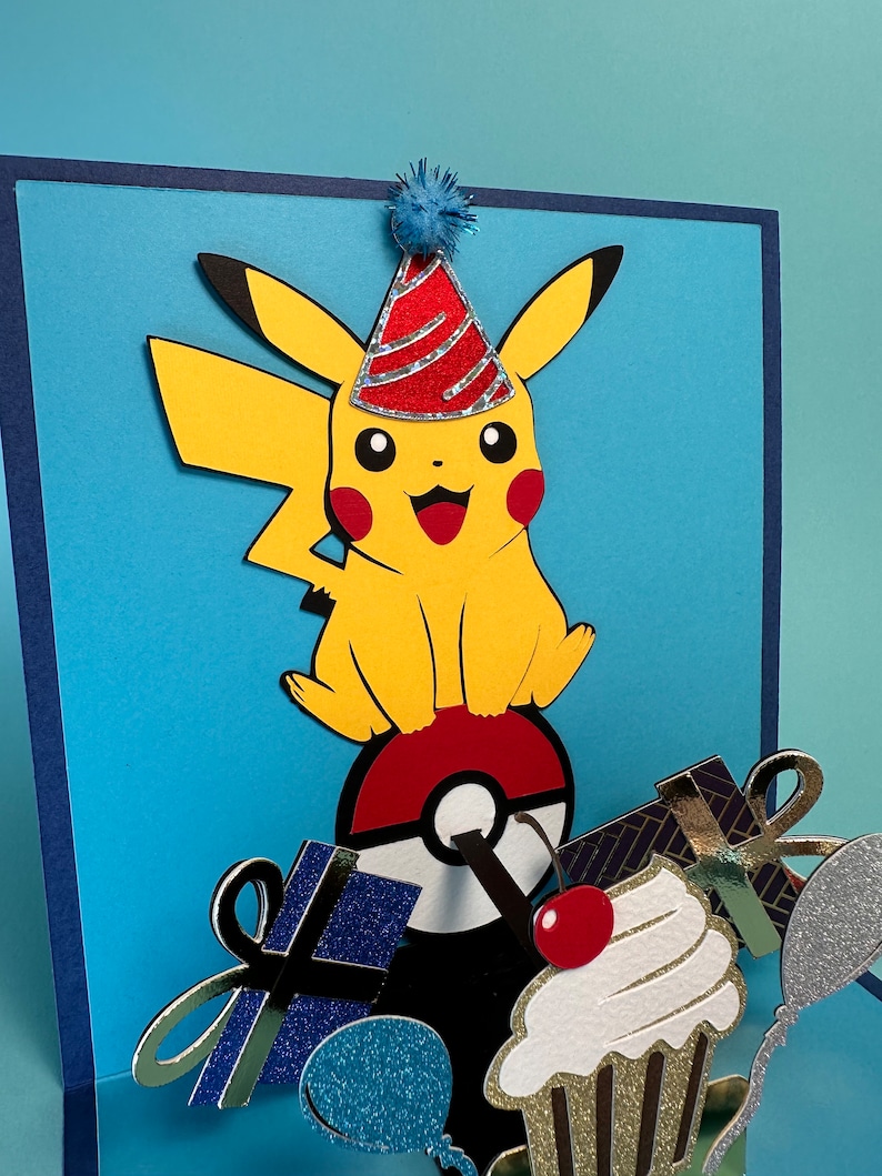 May include: A handmade birthday card featuring a yellow Pikachu character wearing a party hat. The card has a blue background with a pop-up design that includes a Pok&eacute; Ball, gifts, a cupcake, and a balloon.