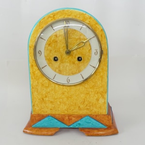 Puede incluir: Un reloj de sobremesa vintage con una esfera texturizada amarilla y una esfera blanca con manecillas doradas. El reloj tiene un borde y una base turquesa con detalles geométricos. El reloj mide aproximadamente 20 cm de alto.