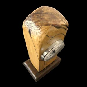 Puede incluir: Un reloj de escritorio de madera con un acabado de veta de madera natural. La esfera del reloj es plateada y redonda, incrustada en el bloque de madera. El reloj se asienta sobre una peque&ntilde;a base de madera marr&oacute;n oscuro. La madera es de color marr&oacute;n claro con acentos marrones m&aacute;s oscuros.
