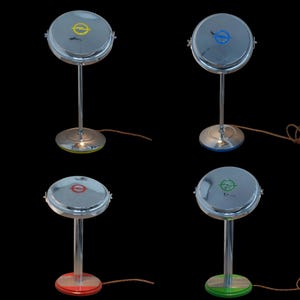 Könnte beinhalten: Vier verchromte Schreibtischlampen mit kreisförmigen Schirmen und Sockeln. Jede Lampe hat eine andere farbige Akzentuierung: gelb, blau, rot und grün. Die Lampen haben ein Retro-Design und einen einzelnen Stiel. Die Lampen stehen auf schwarzem Hintergrund.