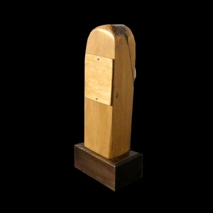 Puede incluir: Una escultura de madera hecha a mano con un acabado marr&oacute;n claro. La escultura tiene una base rectangular y una parte superior alta y redondeada. Un cuadrado de color m&aacute;s claro est&aacute; sujeto al frente con dos tornillos peque&ntilde;os. La escultura est&aacute; sobre un fondo negro.