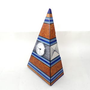 Puede incluir: Un reloj en forma de pirámide con un diseño geométrico marrón, azul y blanco. La esfera del reloj es blanca con números y manecillas negras. La pirámide mide aproximadamente 25 cm de altura.
