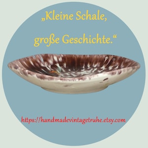 Könnte beinhalten: Eine ovale Vintage-Keramikschale mit braun-weiß gesprenkeltem Innenraum und cremefarbenem Äußeren. Die Schale befindet sich vor einem blauen Hintergrund mit dem Text "Kleine Schale, große Geschichte." und einer Website-Adresse.
