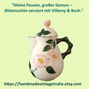 Könnte beinhalten: Eine weiße Keramik-Teekanne mit einem Blumenmuster aus rosa Blüten mit gelben Mitten und grünen Blättern. Der Deckel hat einen Knauf in Form einer rosa Blütenknospe und der Henkel ist dunkelbraun. Text: Kleine Pausen, großer Genuss – Blütenschön serviert mit Villeroy & Boch.