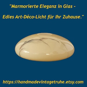Könnte beinhalten: Ein flacher, kuppelförmiger Glaslampenschirm mit cremefarbener Oberfläche und dunkler Marmorierung. Über dem Objekt steht der Text "Marmorierte Eleganz in Glas - Edles Art-Déco-Licht für Ihr Zuhause.", darunter befindet sich eine URL.