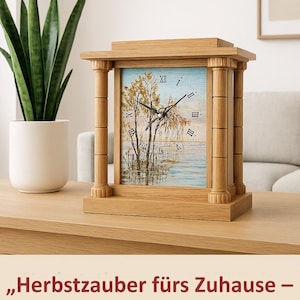 May include: Wooden mantel clock with a landscape painting on the face, featuring a tree and water scene. The clock has Roman numerals and decorative columns. The clock sits on a wooden table next to a potted plant.