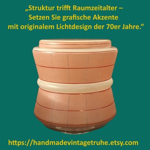 Könnte beinhalten: Ein pfirsichfarbener Glaslampenschirm mit einem Gittermuster aus dünnen roten Linien und einem weißen horizontalen Mittelband. Der Text darüber lautet: Struktur trifft Raumzeitalter – Setzen Sie grafische Akzente mit originalem Lichtdesign der 70er Jahre.