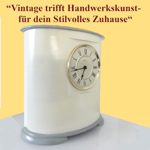 Op de afbeelding: Crèmekleurige vintage klok met een goudkleurige wijzerplaat en zwarte wijzers. De klok heeft een afgeronde rechthoekige vorm met een grijze basis en bovenste rand. De wijzerplaat heeft Romeinse cijfers. Tekst bovenaan luidt: "Vintage ontmoet vakmanschap voor uw stijlvolle huis."