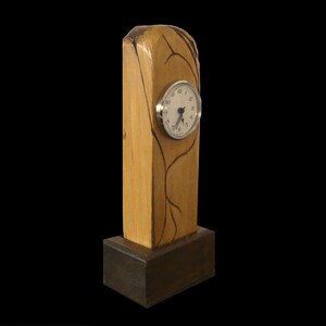 Puede incluir: Un reloj de escritorio de madera con un acabado marr&oacute;n claro y una base marr&oacute;n oscuro. La esfera del reloj es blanca con n&uacute;meros y manecillas negras, en un marco plateado. La madera tiene marcas decorativas oscuras.
