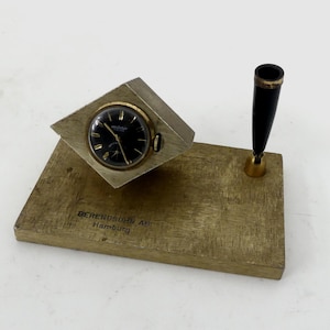 Könnte beinhalten: Ein Vintage-Schreibtisch-Set mit Uhr und Stifthalter. Die Uhr hat ein schwarzes Zifferblatt mit goldenen Akzenten und einer goldfarbenen Lünette. Der Stifthalter hat einen schwarzen Stift mit goldfarbenem Sockel. Die Basis ist rechteckig und trägt den Text "BERENDSOHN Hamburg".