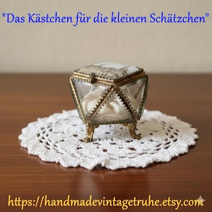 Può includere: Un portagioie vintage di forma geometrica, con pannelli in vetro trasparente e finiture in metallo decorate. La scatola ha un coperchio incernierato con un disegno stampato e poggia su piedini decorativi. Si trova su un centrino bianco all'uncinetto. Il testo dice "Das Kästchen für die kleinen Schätzchen".