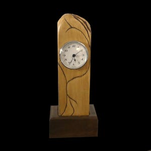 Puede incluir: Un reloj de escritorio de madera con un acabado marr&oacute;n claro y una base marr&oacute;n oscuro. La esfera del reloj es redonda con un bisel plateado y n&uacute;meros negros. L&iacute;neas marrones oscuras que se asemejan a ramas de &aacute;rboles est&aacute;n pintadas en el cuerpo de madera.