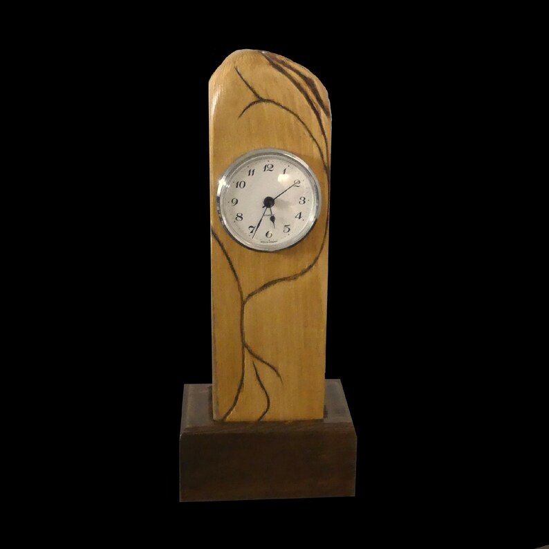 Puede incluir: Un reloj de escritorio de madera con un acabado marr&oacute;n claro y una base marr&oacute;n oscuro. La esfera del reloj es redonda con un bisel plateado y n&uacute;meros negros. L&iacute;neas marrones oscuras que se asemejan a ramas de &aacute;rboles est&aacute;n pintadas en el cuerpo de madera.