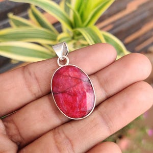 May include: Oval-shaped pendant with a vibrant red gemstone set in a silver-colored metal frame. The gemstone has a natural, textured appearance. The pendant is suspended from a small loop at the top, ready for a chain.