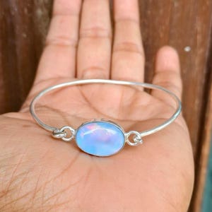 Op de afbeelding: Een zilveren armband met een lichtblauwe ovale opaalsteen. De opaal heeft een parelmoerglans. De armband heeft een eenvoudig, elegant ontwerp. De opaal is ongeveer 2 cm lang.