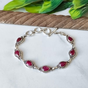 May include: A silver bracelet featuring oval-shaped, deep red gemstones. The bracelet has a toggle clasp and is set on a white surface. The gemstones are evenly spaced along the bracelet's length, creating a symmetrical design.