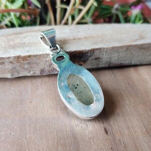 May include: A silver pendant with an oval, light green stone. The stone has dark inclusions and is set in a silver bezel. The pendant has a silver bail for attaching to a chain. The pendant is displayed on a wooden surface.