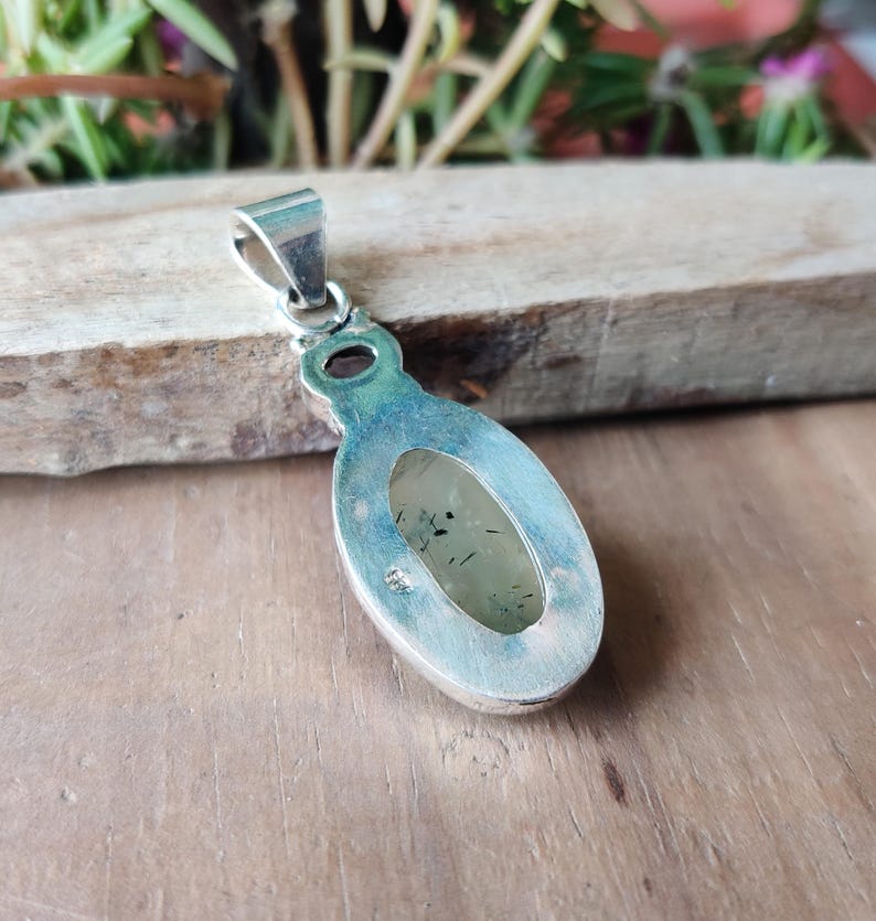May include: A silver pendant with an oval, light green stone. The stone has dark inclusions and is set in a silver bezel. The pendant has a silver bail for attaching to a chain. The pendant is displayed on a wooden surface.