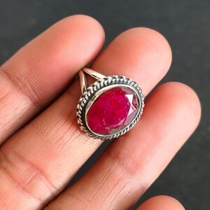 May include: A silver ring with a large, oval-cut ruby gemstone. The ruby is a vibrant red colour and is framed by a decorative, twisted metal border. The ring's band is simple and elegant.