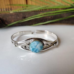 Puede incluir: Una pulsera de plata con una piedra preciosa central redonda de color azul claro. La pulsera presenta un diseño calado decorativo con patrones arremolinados y pequeños acentos redondos. La piedra preciosa tiene un aspecto veteado.